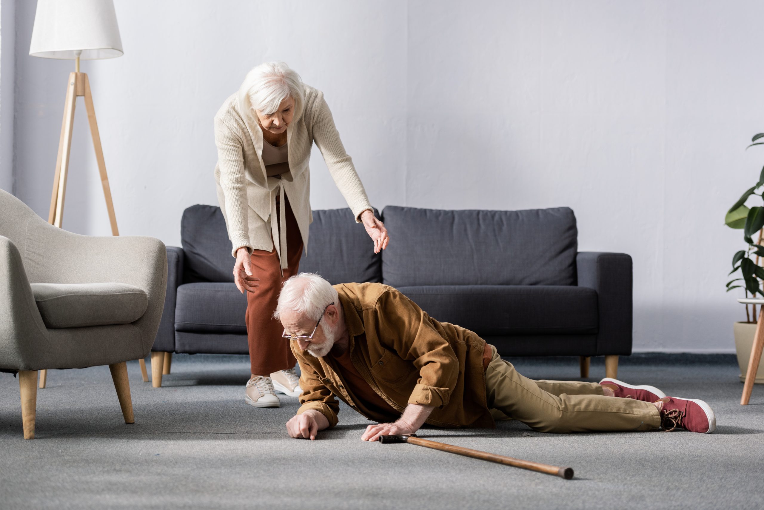 senior woman helping to get up fallen husband lying on floor near walking stick